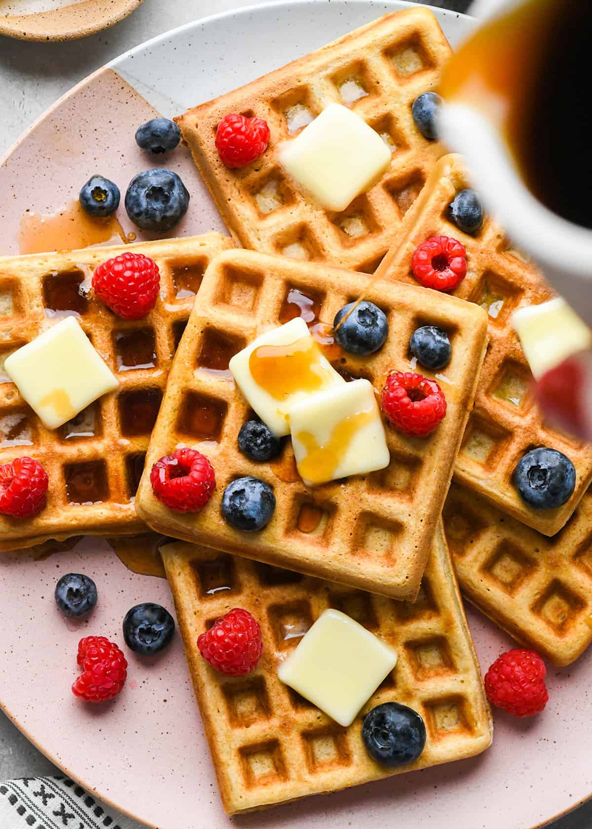 syrup being poured onto 6 Homemade Waffles on a plate topped with butter and berries