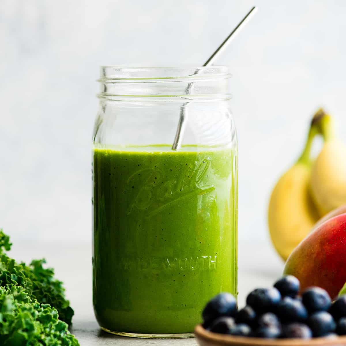 front view of a green smoothie in a large glass mason jar with a metal straw, surrounded with kale, blueberries, bananas, mangoes and green grapes