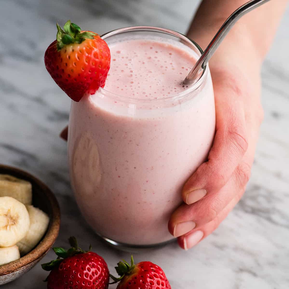 hand holding a glass filled with strawberry banana smoothie with a straw and strawberries.