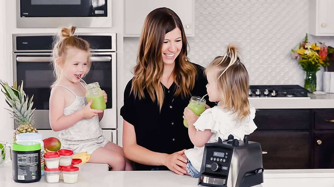 front view of a mom and her two kids sitting on the kitchen counter drinking green smoothies